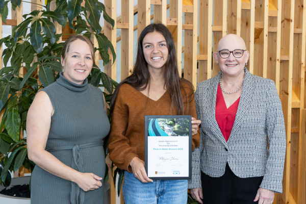 Three women standing in a row with a plant in the background. The woman in the centre is holding a certificate. 